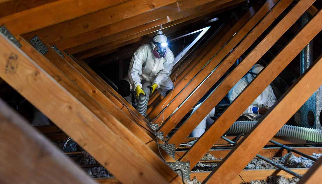 A Home Attics technician in a protective suit and respirator blowing new insulation between the roof beams of an attic